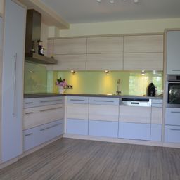 A kitchen with light blue cabinets, a green splashback, and a white vase with flowers. The countertop holds a sink, faucet, and dishwasher.