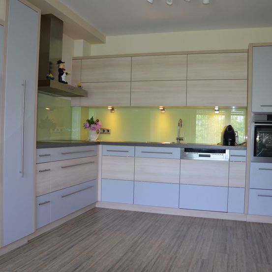 Eine Küche mit hellblauen Schränken, einem grünen Spritzschutz und einer weißen Vase mit Blumen. Die Arbeitsplatte hat ein Waschbecken, einen Wasserhahn und einen Geschirrspüler.