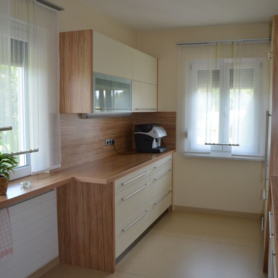 Eine Küche mit Holzschränken, einer Arbeitsplatte und einer Kaffeemaschine. Die Fenster haben weiße Vorhänge.