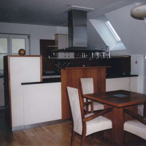 Eine Küche mit Holzschränken, einer Arbeitsplatte und einem Esstisch mit Stühlen. Ein offenes Fenster lässt natürliches Licht herein.