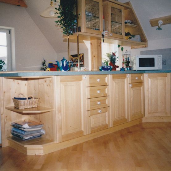 Eine Holzküche mit blauer Arbeitsplatte, Holzschränken und Regalen. Eine Mikrowelle, ein Teepott und ein Korb stehen auf der Arbeitsplatte.