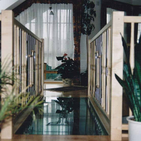 Ein Flur mit Holzgittern, einem Glasboden und Pflanzen im Vordergrund. Dahinter ein Raum mit weißen Vorhängen, einer Topfpflanze und einer Holzbank.