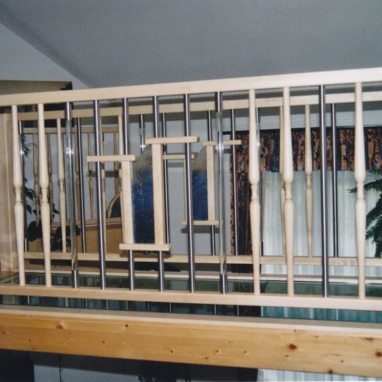 Ein hölzerner Balkon mit Glasboden, Holzgeländer und Metallstangen. Der Balkon hat einen klaren Glasboden, ein Holzgeländer mit vertikalen Stangen und eine Glasscheibe in der Mitte. Der Balkon hat eine weiße Decke.