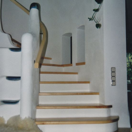 Ein Nahaufnahme einer Holztreppe mit weißen Stufen und hölzernen Handläufen. Die Wand ist weiß, und eine kleine Pflanze ist daran befestigt.