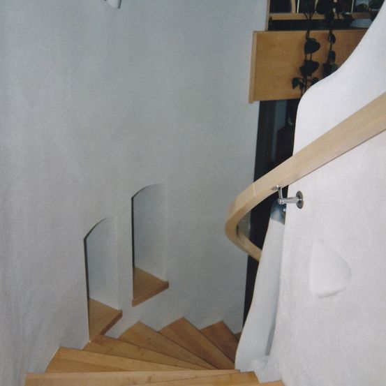 Eine Holztreppe mit weißer Wand, hölzerne Handläufe und eine Pflanze auf einem Holzregal.