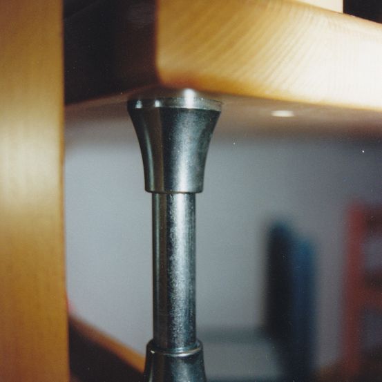 Eine Nahaufnahme eines Metallbeins, das an einem Holzregal befestigt ist, mit einem verschwommenen Hintergrund, der einen Raum zeigt.