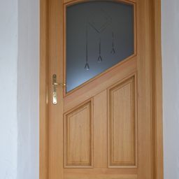 A wooden door with a glass panel features a diamond-shaped design and three hanging objects. The door has a brass handle and is mounted on a white wall.