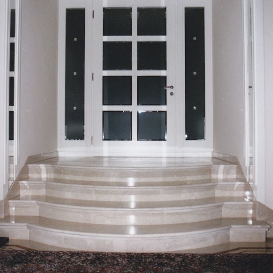 Ein Bild eines modernen Eingangs mit einer großen weißen Tür, die mehrere Glasscheiben aufweist und eine Marmortreppe, die hinaufführt. Die Wände sind weiß und unten befindet sich ein Teppich.