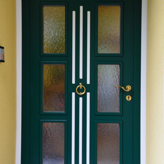 Ein Nahaufnahme einer doppelflügeligen grünen Tür mit Glaseinsätzen und einem Messinggriff. Die Tür hat einen weißen Rand und ist an einer gelben Wand montiert.