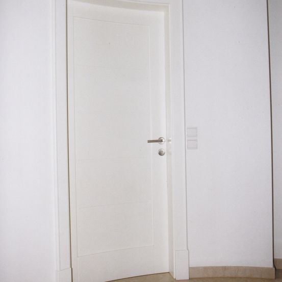 Eine weiße Tür mit silbernem Griff ist geschlossen. Die Tür ist an einer weißen Wand montiert. Es gibt Schalter an der Wand. Der Boden ist gefliest.