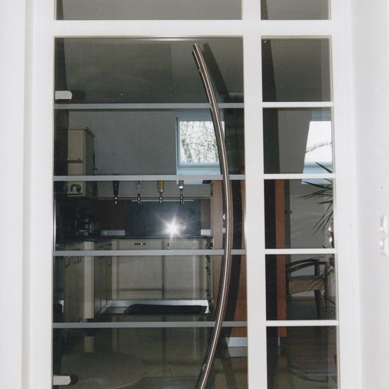 Ein Blick durch eine moderne Glastür zeigt eine Küche mit weißen Schränken, einem Fenster und einem Stuhl mit einer Pflanze. Die Küche hat einen schwarzen Teppich und die Tür einen silbernen Griff.