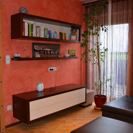 Ein modernes Wohnzimmer mit orangefarbenen Wänden, mit Regalen mit Büchern und einem Bilderrahmen, einem Holzschrank mit Schubladen und einer hohen Topfpflanze.