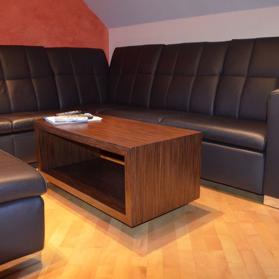 Ein Wohnzimmer mit einem dunklen Leder-L-Sofa und einem hölzernen Couchtisch davor, mit Fernbedienungen und Büchern darauf. Der Raum hat Holzböden und eine orange Wand.