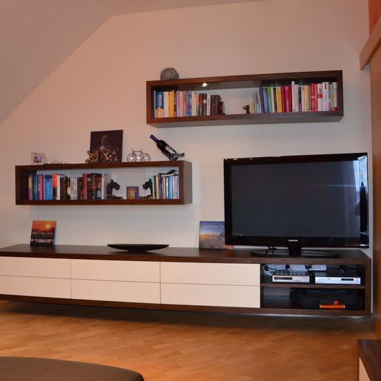 Ein modernes Wohnzimmer mit Holzboden zeigt einen Fernseher auf einem Holzschrank unter zwei schwebenden Regalen, eines mit Büchern und das andere mit einer Flasche, einem Bild und einem Fahrrad.