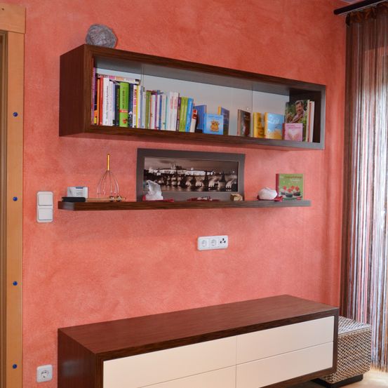 Ein Raum mit pfirsichfarbener Wand, zwei Holzregalen mit Büchern und einem gerahmten Bild auf dem oberen Regal. Unten steht ein Schrank mit Schubladen.