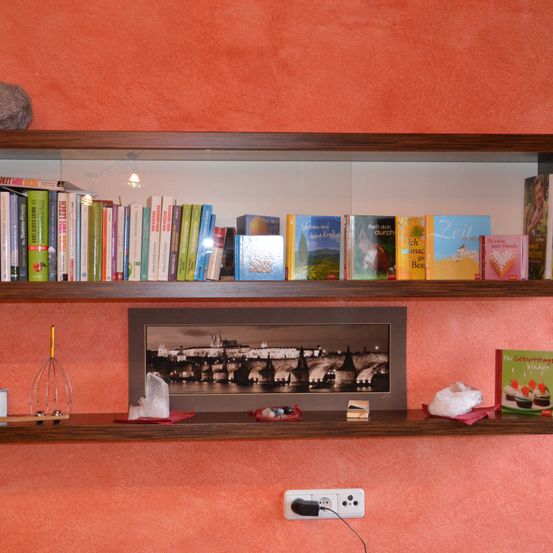 Ein hölzernes Regal mit zwei Ebenen, das Bücher, ein gerahmtes Foto einer Burg und verschiedene Gegenstände zeigt. Eine Steckdose ist an der Wand sichtbar.