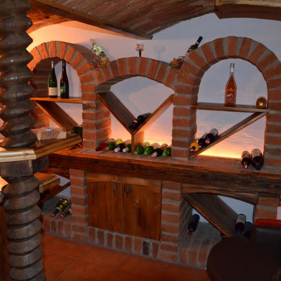 Ein Weinkeller aus Ziegelsteinen mit gewölbten Regalen, beleuchtet von gelbem Licht. Flaschen Wein sind auf den Regalen ausgestellt. Ein hölzerner Tresen hält zusätzliche Flaschen.