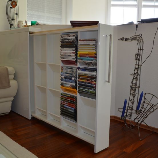 Ein weißer Bücherregal gefüllt mit CDs und DVDs in einem Raum mit einer Couch, einem Fenster und einem SAXOFON-förmigen Ständer an der Wand.