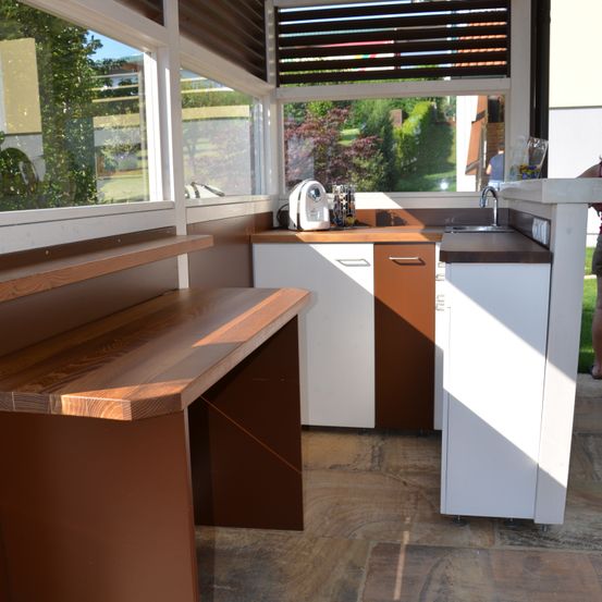 Eine Außenküche mit Holzarbeitsplatten, weißen Schränken und einem Waschbecken. Der Bereich ist durch Glasfenster abgeschlossen und verfügt über eine Kaffeemaschine auf der Arbeitsplatte.