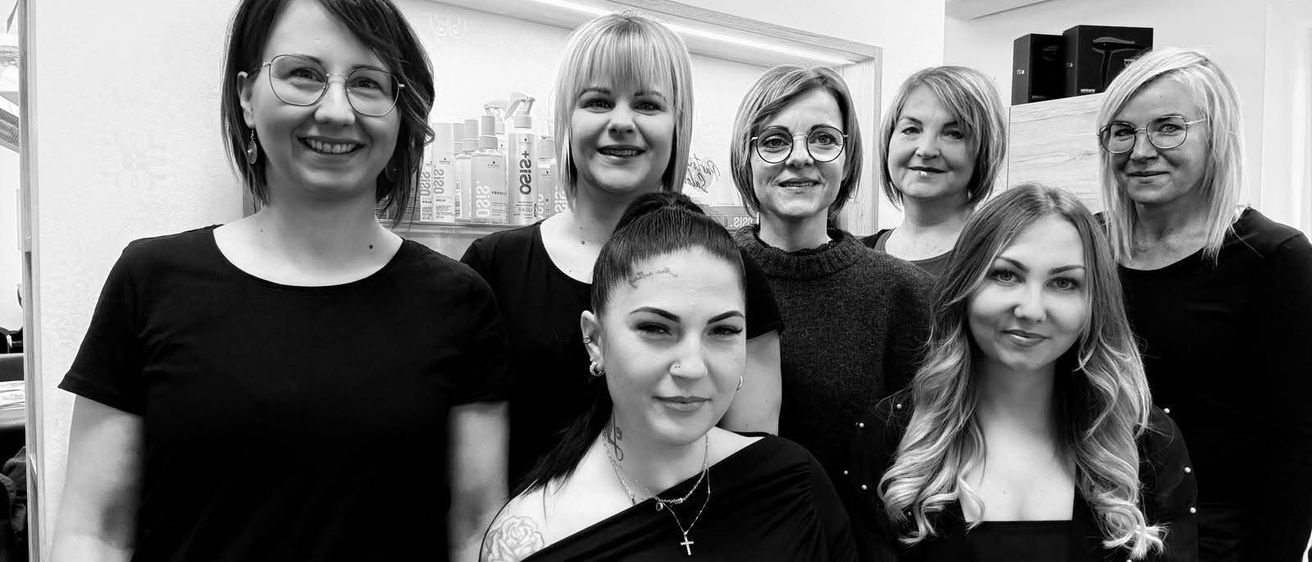 Eine Gruppe von Frauen in schwarzer Kleidung, darunter eine mit Tätowierung und Halskette, posiert für ein Foto in einem Raum mit weißer Wand und einem Regal mit Flaschen.