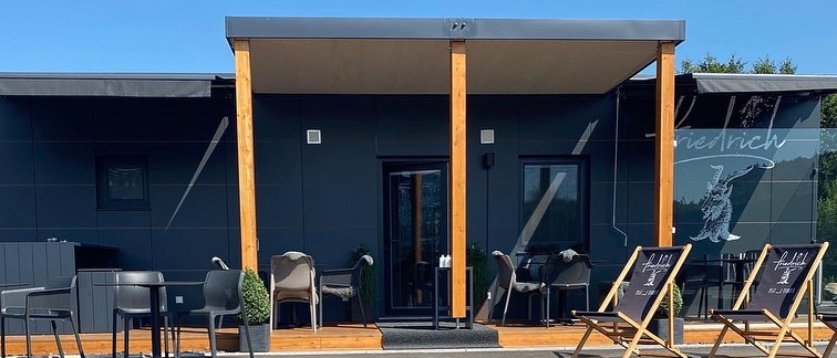 Ein modernes, schwarzes Gebäude mit Holzpfosten hat eine überdachte Veranda. Draußen sind Tische und Stühle aufgestellt. Zwei Holzstühle haben Schilder darauf.