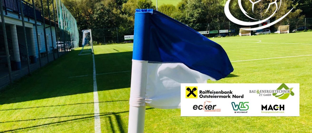 Ein Fußballfeld mit einer blau-weißen Flagge auf einem weißen Pfosten, umgeben von einem grünen Feld und Bäumen. Im Hintergrund befindet sich ein Fußballlogo und ein Banner mit Sponsoren.
