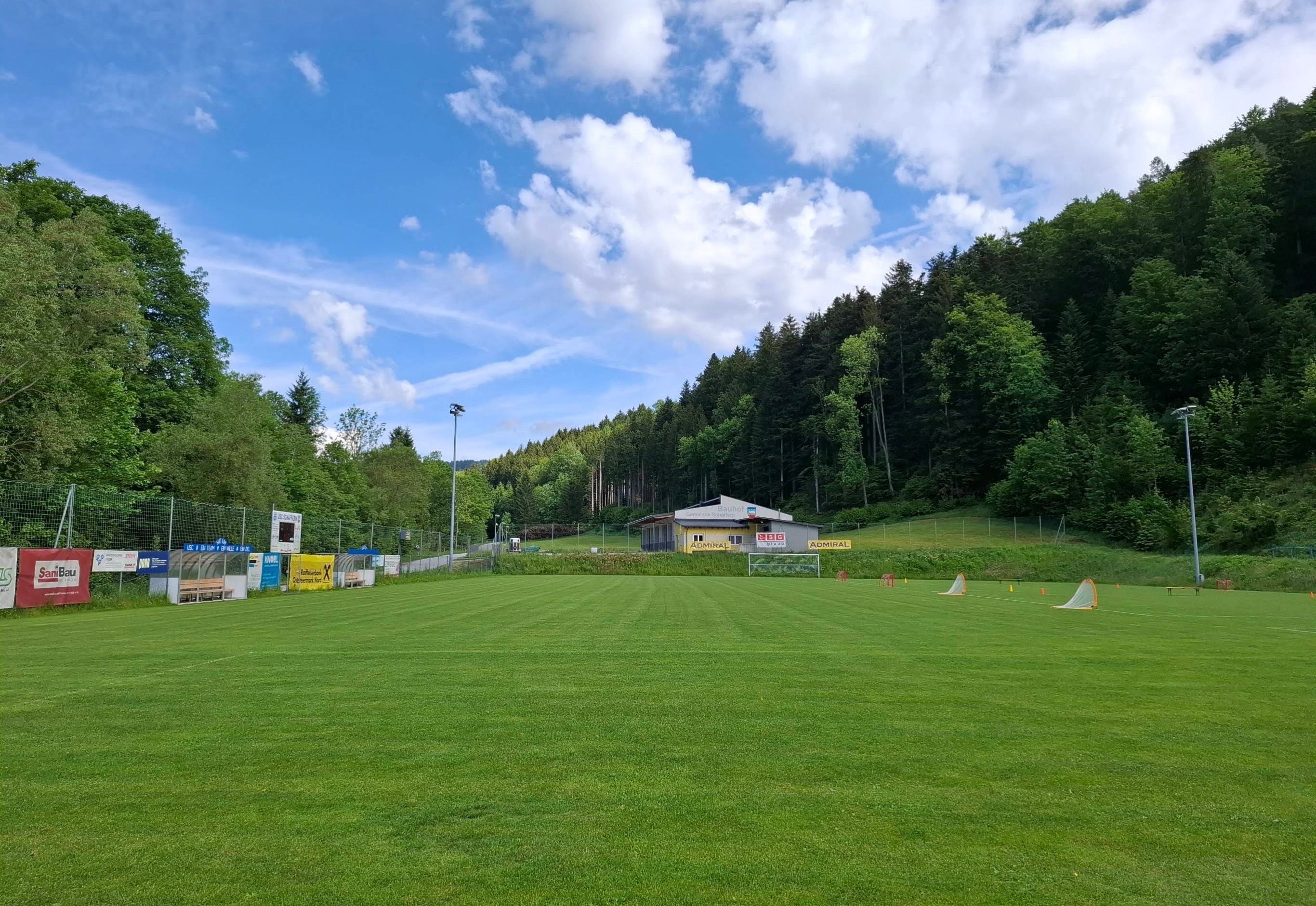 Ein leeres Fußballfeld mit einem kleinen Gebäude und einer Anzeigetafel im Hintergrund. Das Feld ist von einem Zaun und Bäumen umgeben. Der Himmel ist blau mit einigen Wolken.