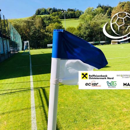 Ein Fußballfeld mit einer blau-weißen Flagge auf einem weißen Pfosten, umgeben von grünem Rasen und einer weißen Linie. Im Hintergrund befinden sich ein Fußballlogo, Bäume und ein Gebäude.