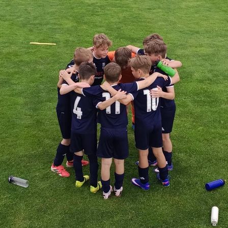 Eine Gruppe junger Fußballer in marineblauen Trikots steht auf einem Rasenfeld versammelt, wahrscheinlich in Vorbereitung auf ein Spiel. Sie haben Nummern auf dem Rücken, und zwei Wasserflaschen liegen auf dem Boden.