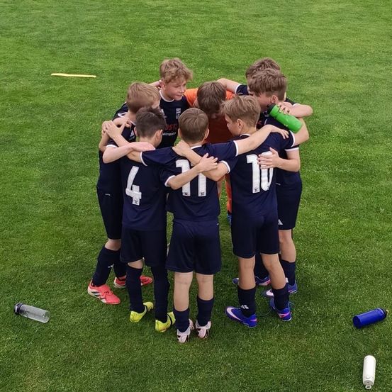 Eine Gruppe junger Fußballer in marineblauen Trikots steht auf einem Rasenfeld versammelt, wahrscheinlich in Vorbereitung auf ein Spiel. Sie haben Nummern auf dem Rücken, und zwei Wasserflaschen liegen auf dem Boden.