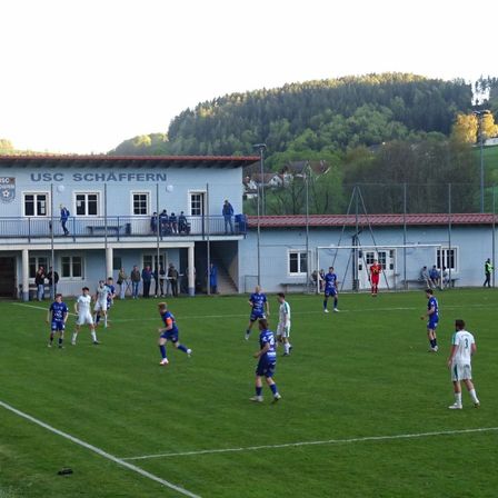 Ein Fußballspiel ist auf einem Feld im Gange, mit einem blauen Gebäude, das USC Schaffern im Hintergrund trägt. Spieler in blauen und weißen Uniformen sind aktiv an dem Spiel beteiligt, während andere an den Seitenlinien stehen. Das Feld ist gut gepflegt, und die Umgebung umfasst Bäume und einen Zaun.