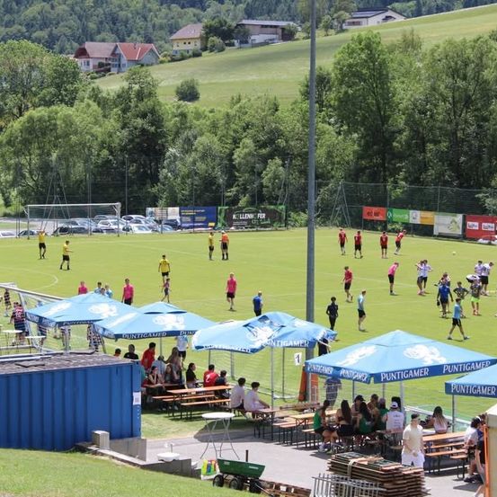 Ein Fußballfeld mit Spielern in verschiedenen Trikots verstreut. Zuschauer sitzen unter blauen Schirmen und beobachten das Spiel. Im Hintergrund sind Häuser und Bäume zu sehen.