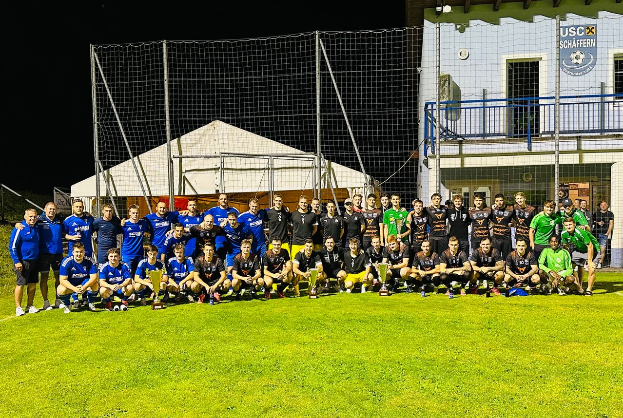 Eine Fußballmannschaft steht auf einem Feld mit Trophäen und trägt Trikots mit verschiedenen Nummern und Farben. Im Hintergrund befindet sich ein Gebäude mit Balkon.