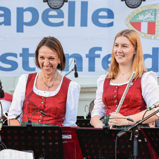 Zwei Frauen in traditioneller bayerischer Kleidung stehen hinter Notenständern, lächelnd und halten Musikinstrumente. Ein Banner mit blauem und weißem Text ist im Hintergrund.