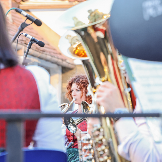 Eine Frau mit rötlichen Locken spielt ein Blechblasinstrument während eines Konzerts. Die Szene ist im Freien.