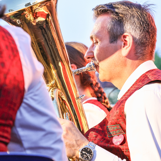 Ein Mann in einem roten Weste spielt eine Messing-Tuba, mit einer anderen Person in Rot neben ihm und einem unscharfen Hintergrund.