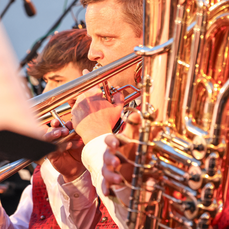 Ein Mann spielt die Posaune, während ein anderer Musiker hinter ihm teilweise verschwommen ist. Das Instrument hat einen glänzenden, goldenen Finish und der Mann ist in einer roten Weste gekleidet.