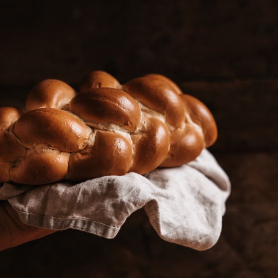 Eine Person hält ein goldbraunes geflochtenes Brot auf einem weißen Tuch vor einem dunklen Hintergrund.