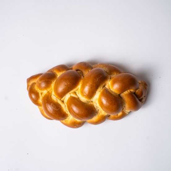 Ein Laib traditionelles Challah-Brot ist auf einer weißen Oberfläche zu sehen. Das Brot hat eine goldbraune Farbe und ist aufwendig geflochten. Es hat eine leicht erhöhte Textur, was auf ein frisch gebackenes Brot hinweist.