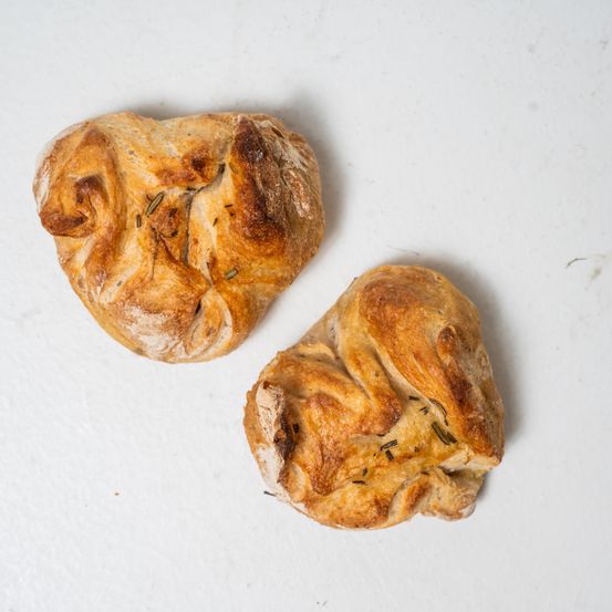 Zwei herzförmige Brote mit sichtbaren Rosmarinblättern auf einer weißen Oberfläche.