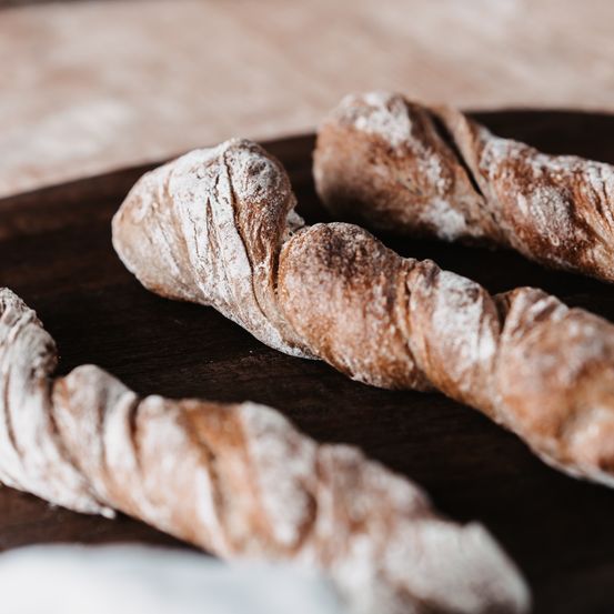 Drei frisch gebackene Brotlaibe mit knuspriger Kruste und leichter Mehlbestäubung liegen auf einem Holzbrett.