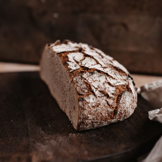 Ein Stück Vollkornbrot mit rauem Äußeren liegt auf einem Holzbrett. Die Kruste ist leicht angebrannt und die Oberfläche ist mit Mehl bestreut.