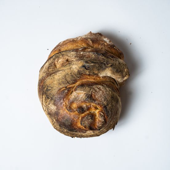 Ein Laib handwerklich gebackenes Brot mit einer rauen, gebrannten Kruste, die auf einer weißen Oberfläche ruht. Das Brot ist leicht verdreht, was ihm eine einzigartige Form verleiht.