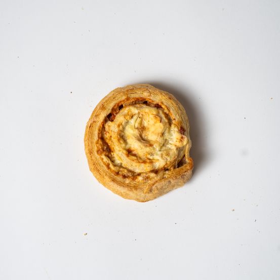 Ein Gebäck, möglicherweise ein Käsebrötchen, wird auf einer weißen Oberfläche präsentiert. Es hat ein Spiraldesign und eine goldbraune Kruste.