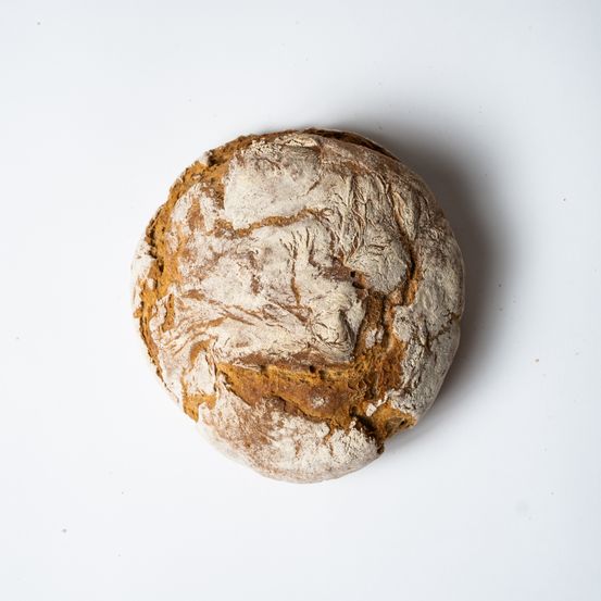 Ein runder Laib Brot mit rustikalem Aussehen, golden-brauner Kruste und leicht ungleichmäßiger Oberfläche, auf einem schlichten weißen Hintergrund liegend.
