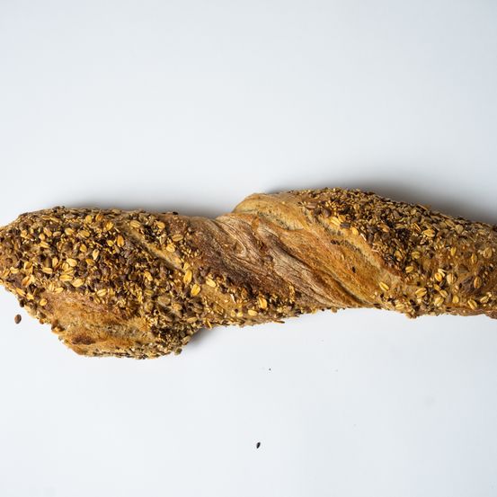 Ein getwistetes Brot mit knuspriger, körnerreicher Kruste liegt auf einer weißen Oberfläche, möglicherweise einem Tisch. Das Brot hat eine braune, leicht verbrannte Farbe und ist mit Samen bestreut.