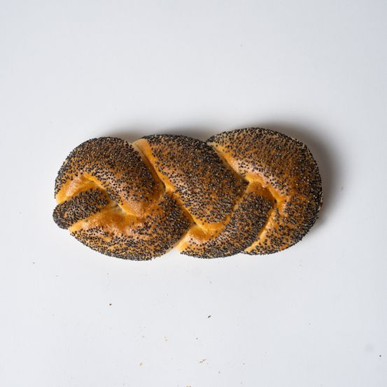 Ein gebackenes Brötchen mit Mohn liegt auf einer weißen Oberfläche. Der Teig hat eine goldbraune Farbe und ist mit schwarzen Mohnsamen verziert.