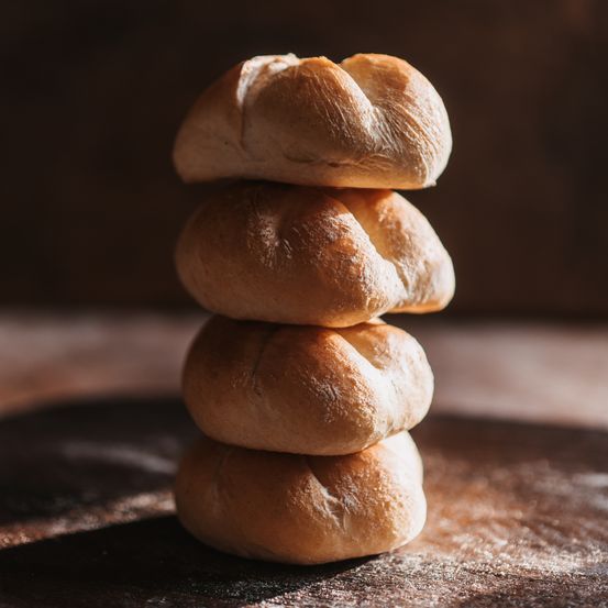 Vier Brötchen sind aufeinandergestapelt. Sie sind goldbraun und scheinen frisch gebacken zu sein. Der Hintergrund ist dunkel, möglicherweise eine Holzoberfläche.