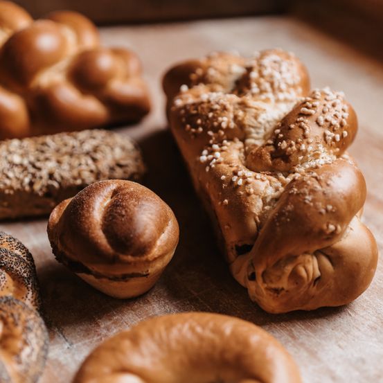 Ein Holzbrett zeigt verschiedene Brotsorten, darunter ein geflochtener Laib mit Sesamkörnern, ein rundes Gebäck und ein Laib mit Mohn.
