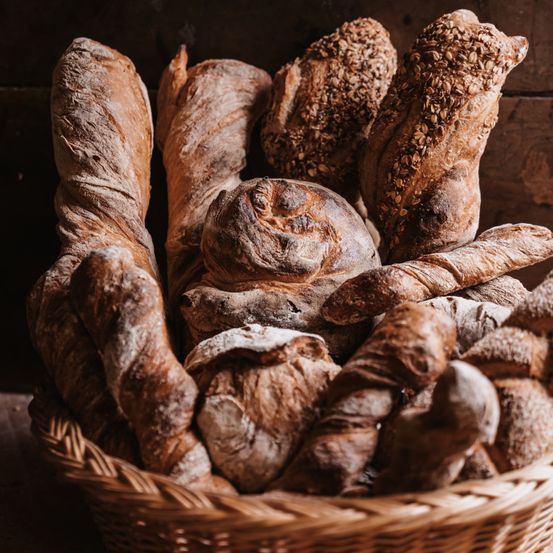 Ein Korb aus Weide gefüllt mit verschiedenen Brotsorten, darunter Baguettes und Laibe, präsentiert auf einem hölzernen Hintergrund.